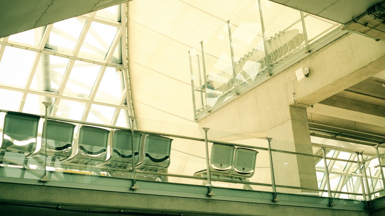 Rows of seats in an airport waiting area beneath a glass-walled hall